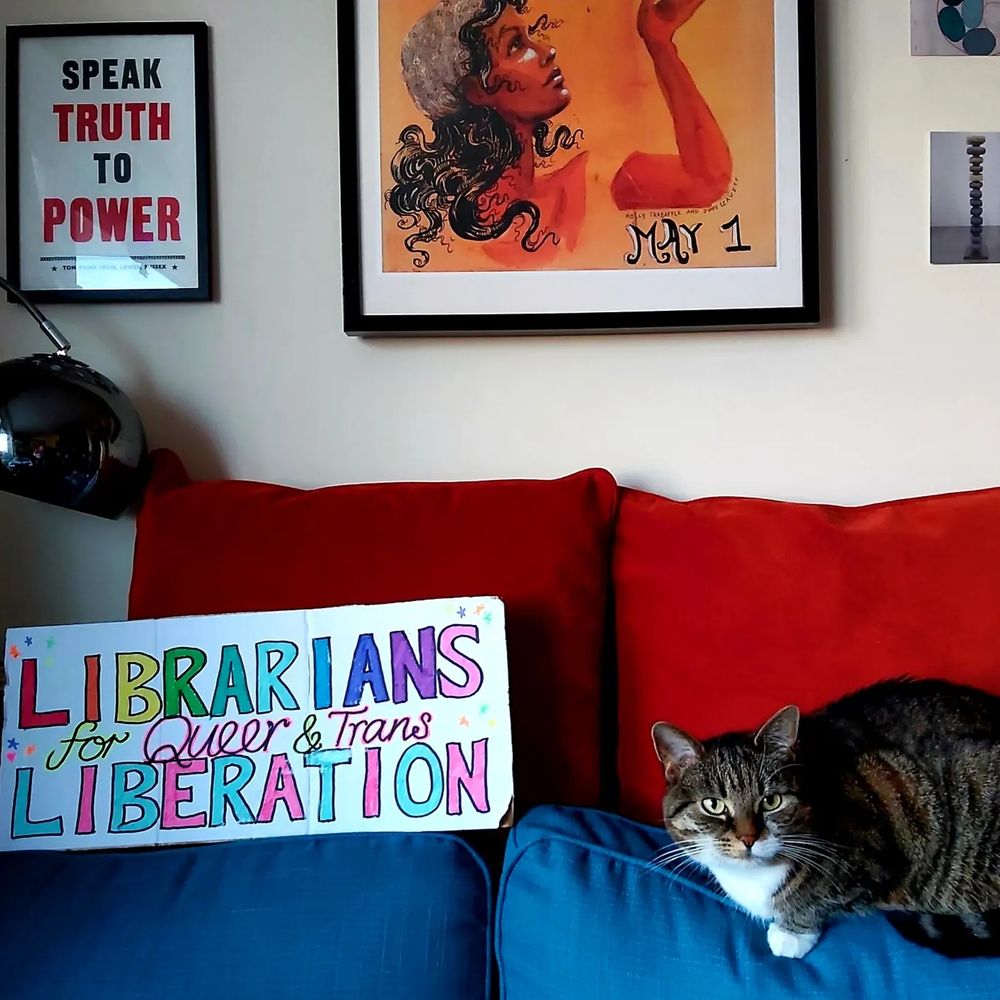 A colourful hand-drawn placard reading LIBRARIANS for Queer and Trans LIBERATION next to a tabby cat on a sofa facing the camera. Above them are some framed graphic prints reading SPEAK TRUTH TO POWER and May 1. 