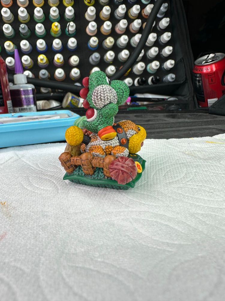 A small 3-4” resin 3d printed model of Yoshi, from Nintendo’s Mario Bros series, sitting atop his companion Poochy.  Much of the figure looks like it crocheted using sculpted details.  It is painted using bright saturated colors and is placed on a paper towel in front of a paint organizer.  There are two empty Coke Zero cans and a superglue bottle in the background.