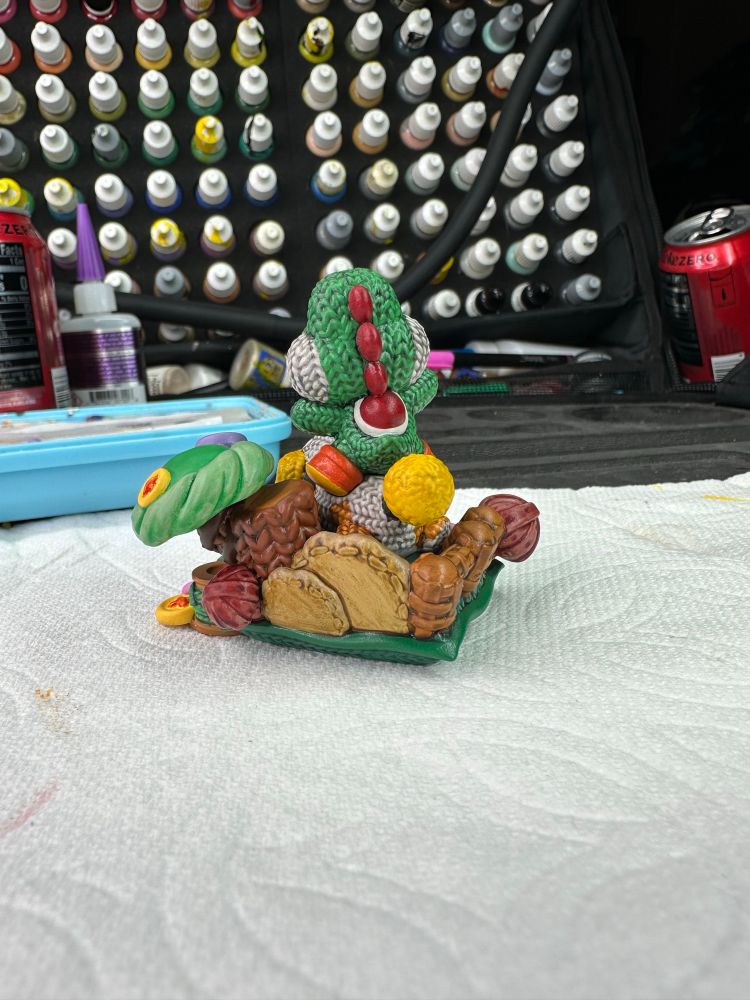 A small 3-4” resin 3d printed model of Yoshi, from Nintendo’s Mario Bros series, sitting atop his companion Poochy.  Much of the figure looks like it crocheted using sculpted details.  It is painted using bright saturated colors and is placed on a paper towel in front of a paint organizer.  There are two empty Coke Zero cans and a superglue bottle in the background.