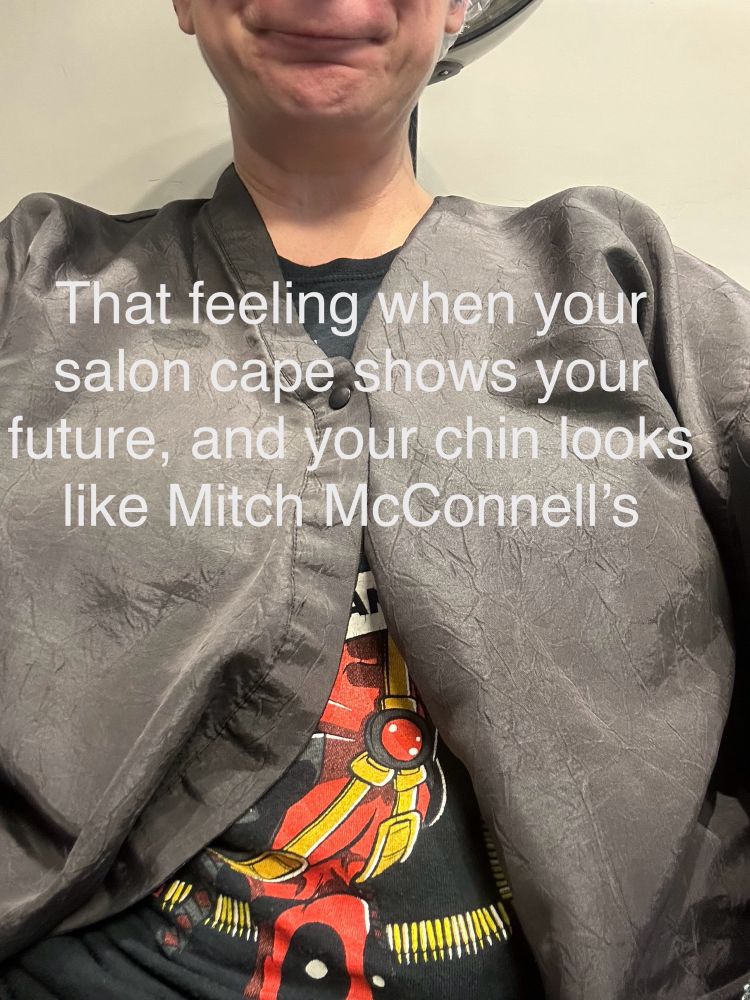 A person sits with a protective grey apron cape draping asymmetrically as if boob, and a very short chin that looks like a turtle