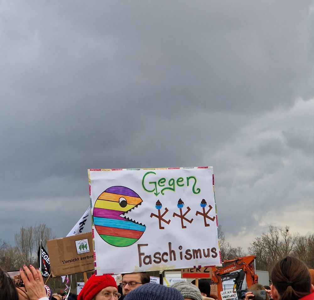 Ein weißes Plakat, mit einem Regenbogen-PacMan mit offenem Mund links auf dem Plakat.
Rechts davon braune Halenkreuz-Männchen mit blauen Gesichtern und einem braunen Haufen auf dem Kopf.