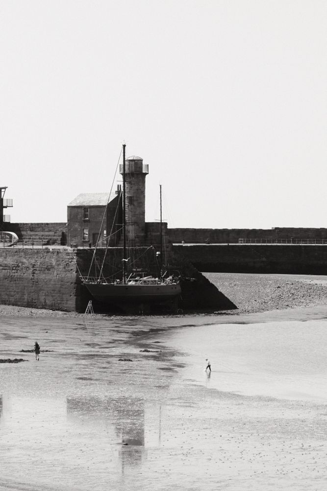 Two people walk on the beach behind a harbour wall. A boat is being repaired up against the harbour wall