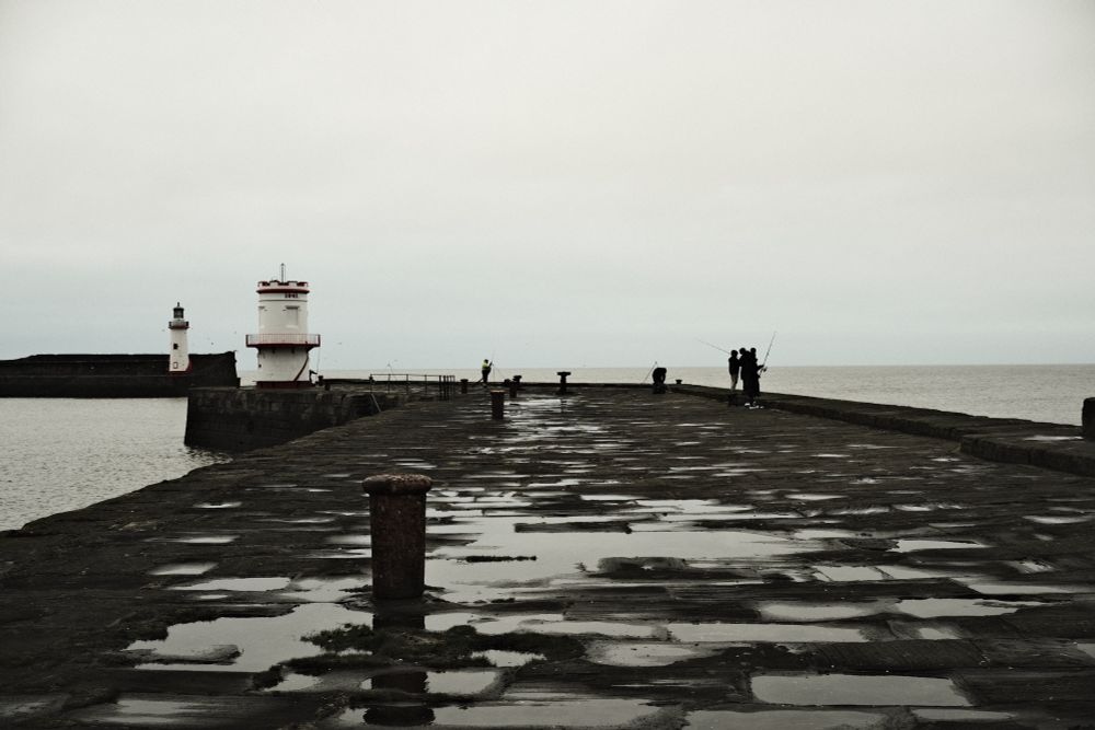 A view down the harbour wall. There are several people fishing off the edge of the walls and in the background are the lighthouse towers to show the ends of the two harbour walls