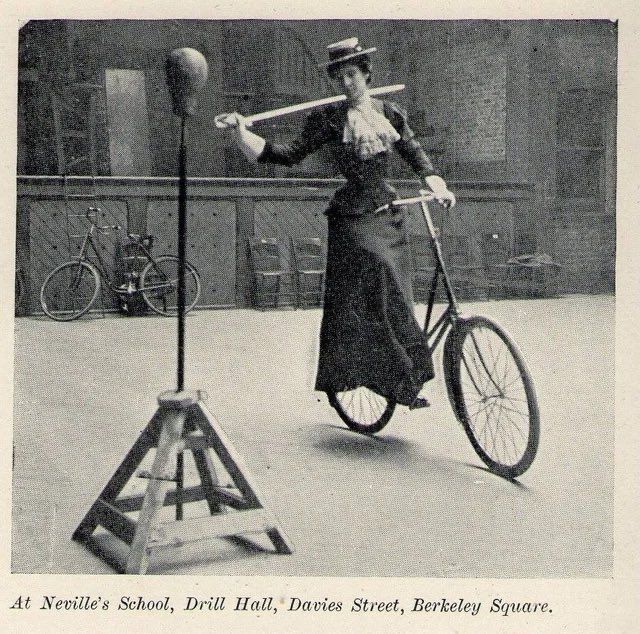 Foto sèpia on es veu una dona, enfundada en un vestit de principies de segle, muntant en bicicleta i amb un sabre a la mà. Va rondat i està punt de fotre-li una bona estocada a una mena de "punch ball".