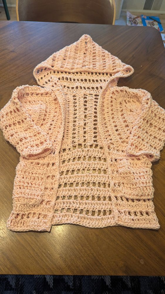 Light pink, hooded crocheted cardigan spread out on a wood table. It's pretty much all in double crochet, in alternating rows of all DC and DC, ch-1, DC. It's one of the hexagon cardigans from Make and Do Crew and the yarn is Lion Brand, Feels Like Butta. 