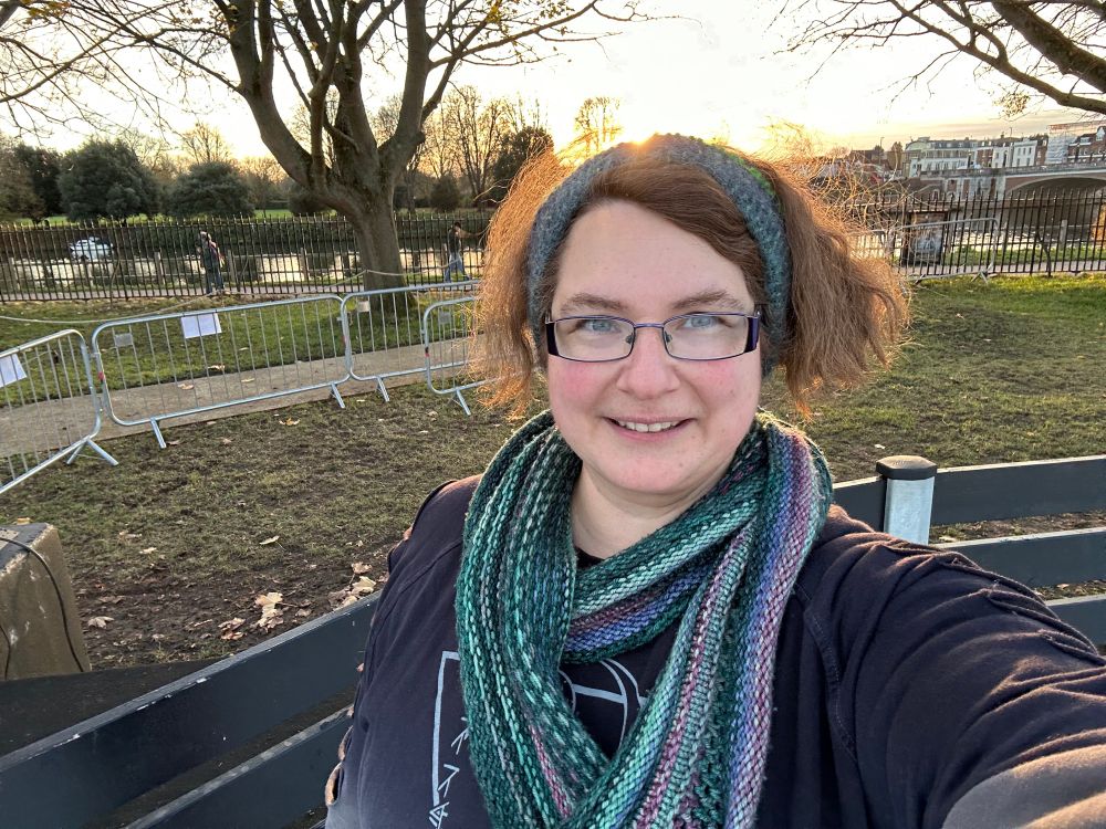 A selfie of me looking pink cheeked and cold on an ice rink in front of the Thames (same ice rink, facing the other way).