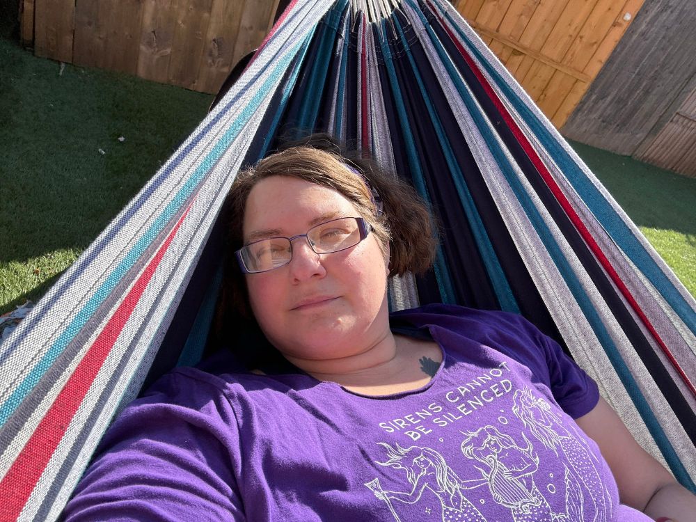 Me in a hammock! Chest up selfie lying in a stripy hammock. My eyes are half closed. I’m in my garden, it’s sunny. Wearing glasses and a purple t-shirt with mermaids on that says “sirens cannot be silenced”.