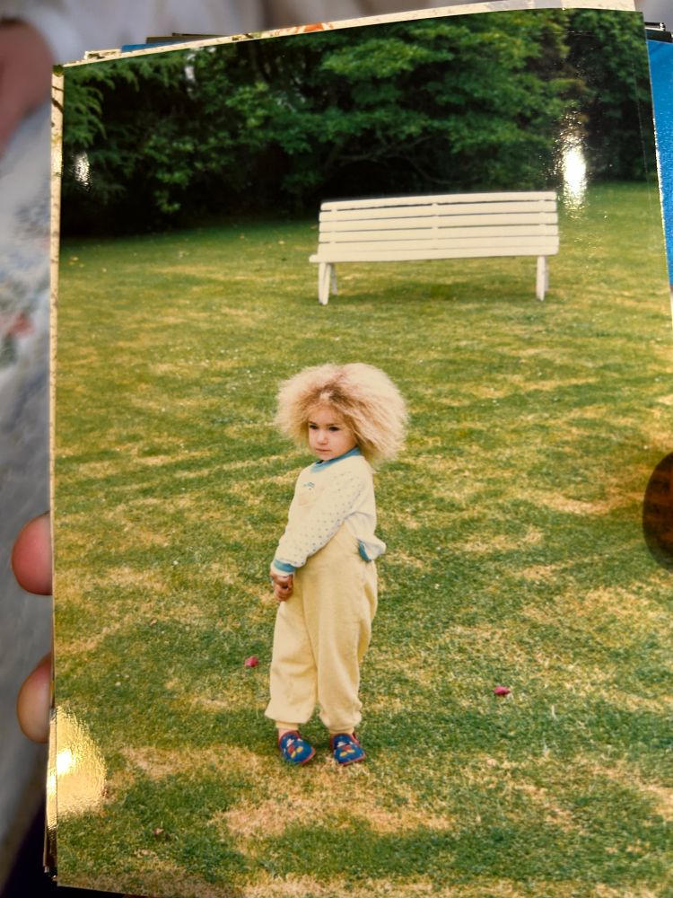 A photograph of a small girl in yellow overalls with a cloud of blond dandelion fuzz hair standing on a lawn looking solemnly off camera.
