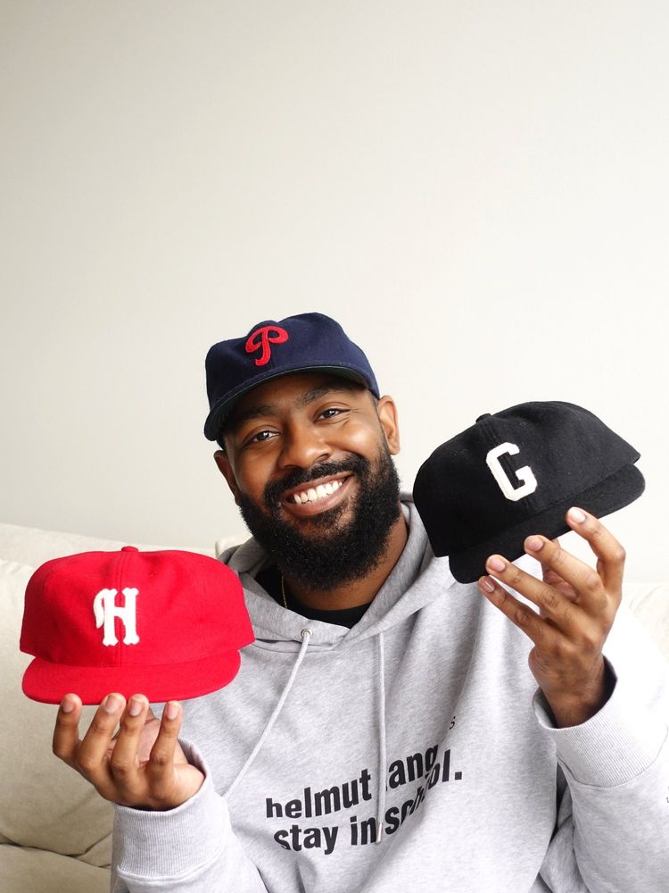 A man showing three Ebbets Field Flannel caps. They have baseball team logos on them. 