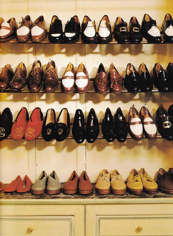 Racks of shoes. Many are spectator shoes made from a combination of brown calf and white buckskin leather. There are some red slippers. Also black shoes with silver buckles. Some of the slippers have intricate monograms at the toe. 