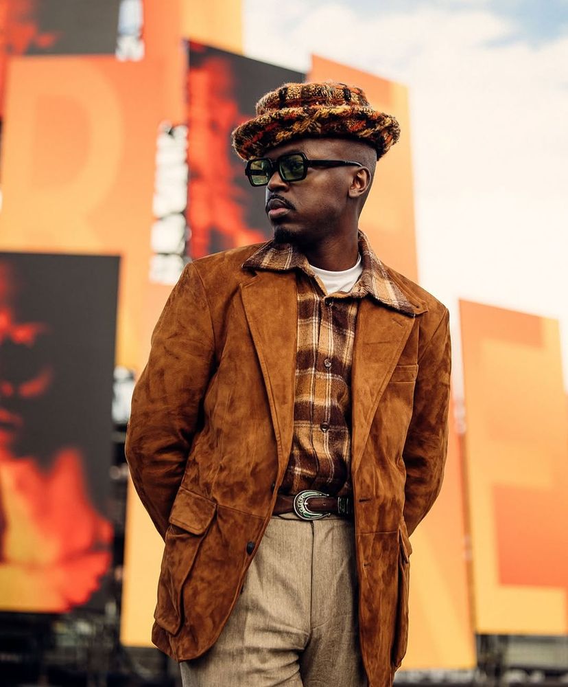 A man wears a brown suede sport coat with tan trousers, brown plaid shirt, and a fuzzy plaid hat. 