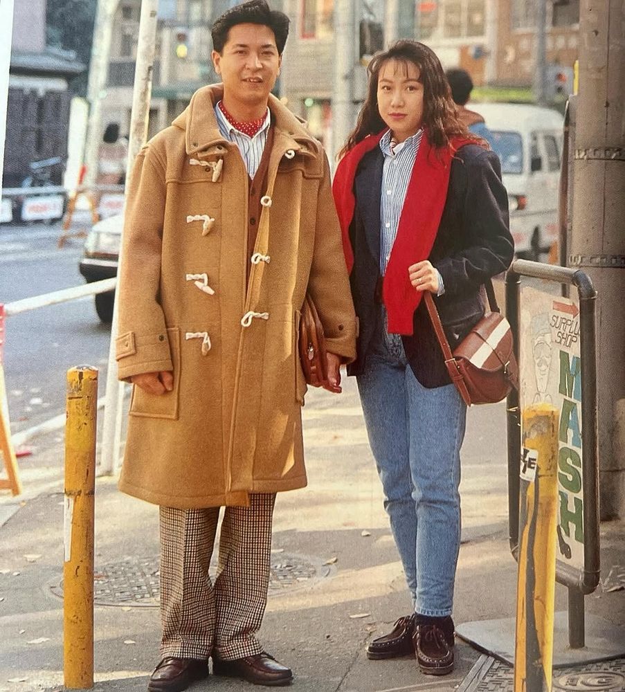Photo of a man and a woman. The man is wearing a tan duffle coat with brown checked trousers. The woman is wearing a navy sport coat with light blue jeans. 