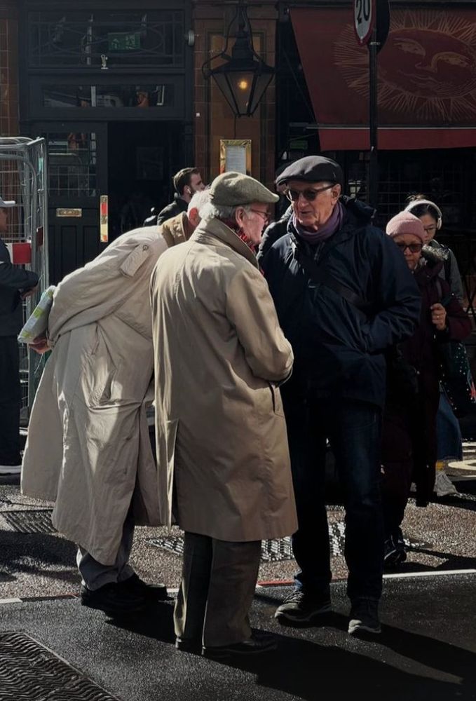 Bunch of older men standing around in big raincoats and tweed flat caps. 