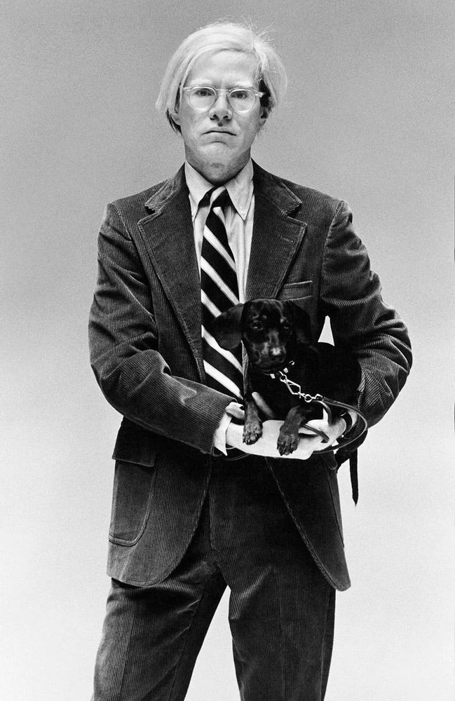Andy Warhol wearing a corduroy single-breasted suit and white shirt. His tie is worn with a four-in-hand knot. He's also carrying a small dog. 