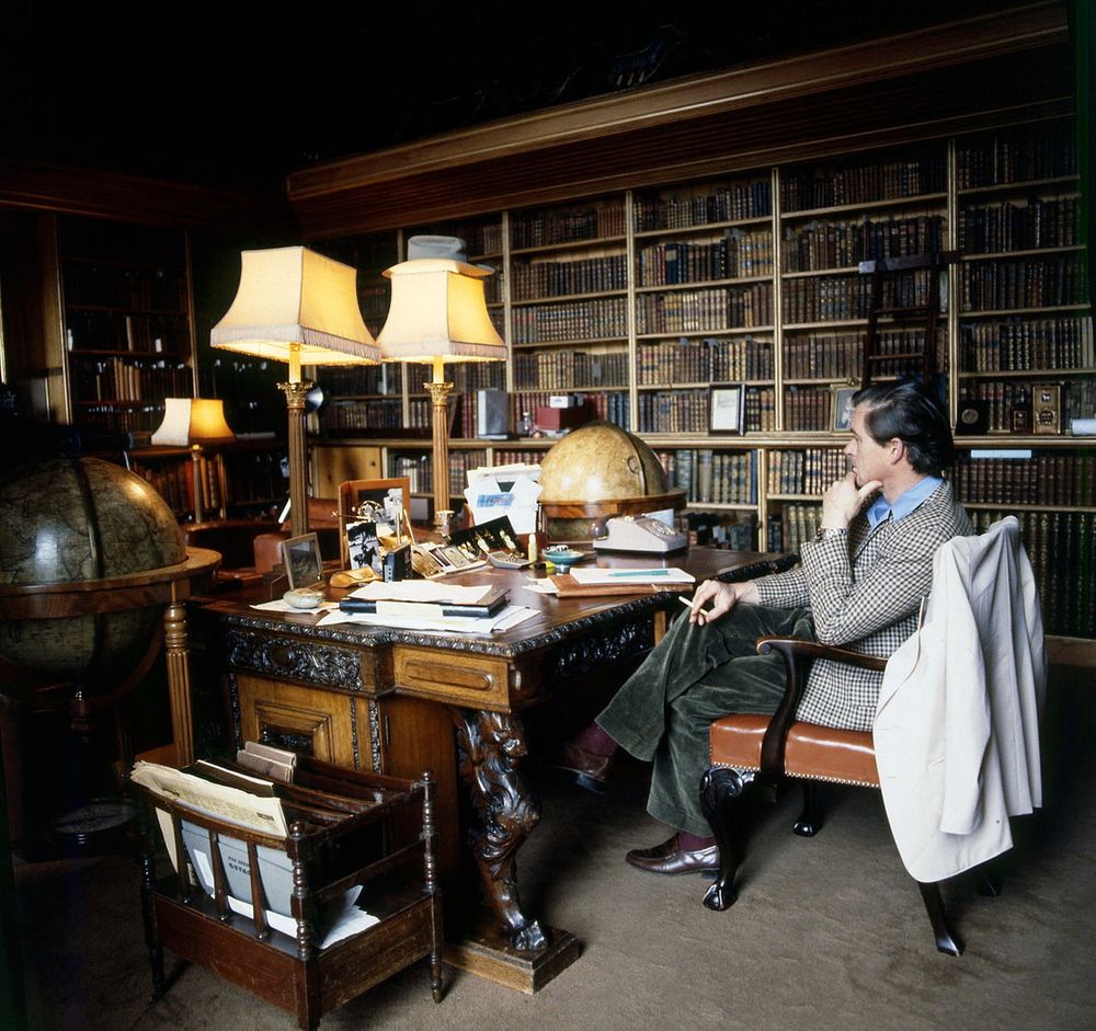 A British duke sitting at home in a checked tweed sport coat and green corduroys. He's surrounded by books, fancy wood desk, and some fancy lamps. 