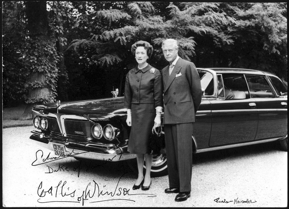 The Duke standing with his wife in front of a car. He's wearing a dark double-breasted suit with a white shirt, a pocket square, and a tie with a big knot. 
