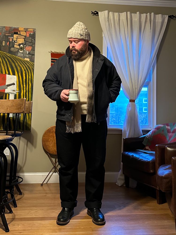 A man wearing a navy deck jacket with black jeans, black boots, cream turtleneck, grey scarf, and grey beanie