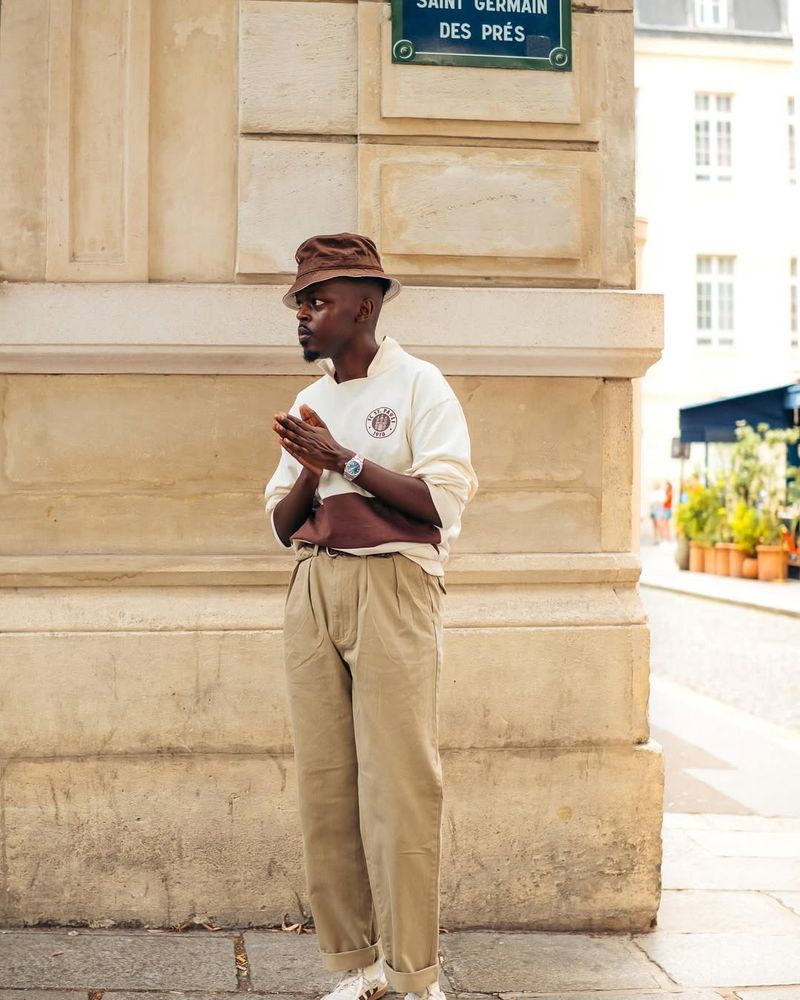 A man wears a pullover knit with tan chinos and a brown bucket hat. 