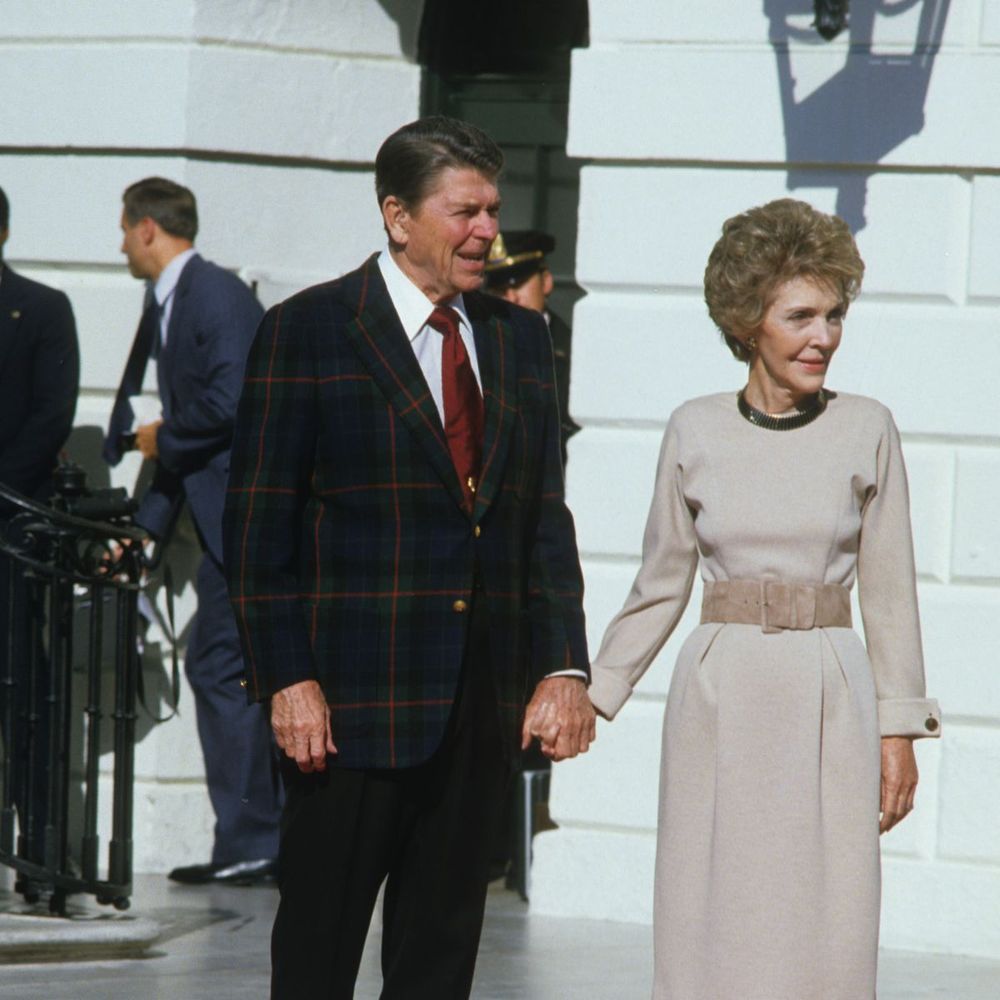 Reagan in a green tartan blazer, charcoal trousers, white shirt, and red tie. 