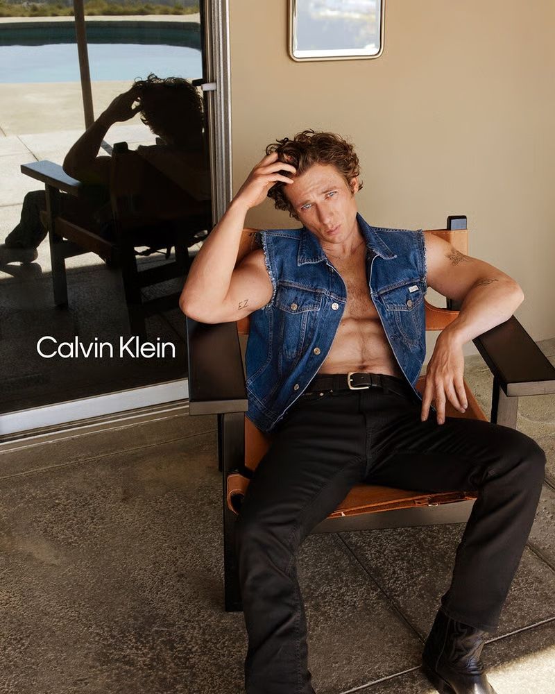 Jeremy Allen White in a Calvin Klein ad wearing black jeans, black boot, and a blue denim vest. 