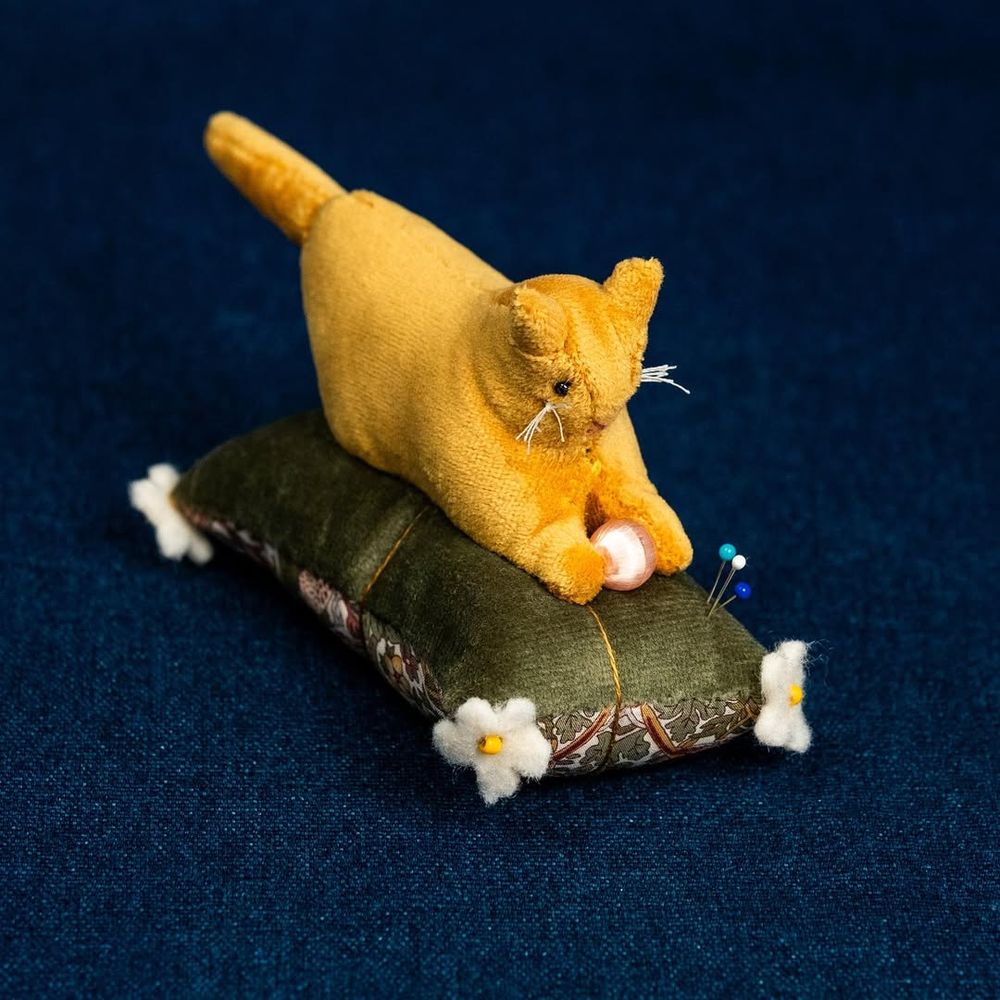 Another cat-shaped pin cushion. There is a yellow colored cat sitting on a green pillow, which holds some pins. 