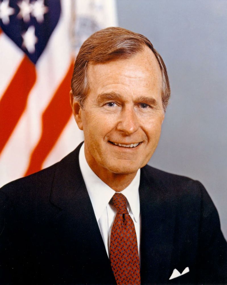 Bush wearing a navy suit with a white shirt and white pocket square. HIs red tie is tied in a four-in-hand. 