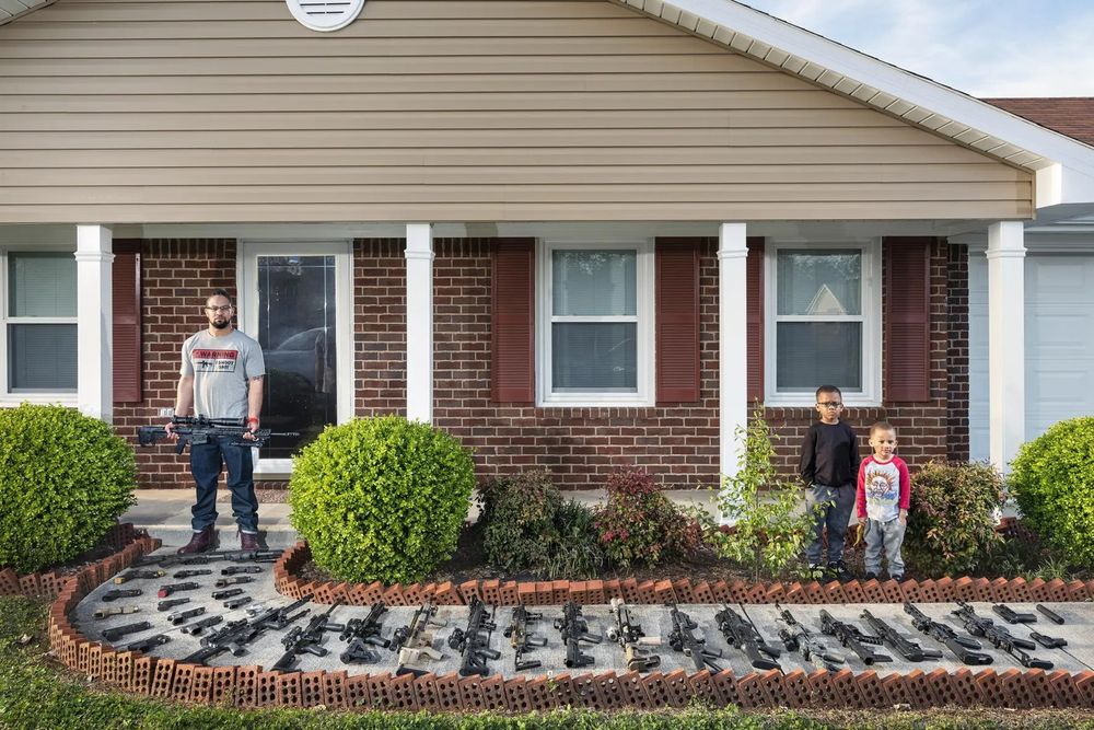 A man outside his home. The walkway into his home is full of guns. 