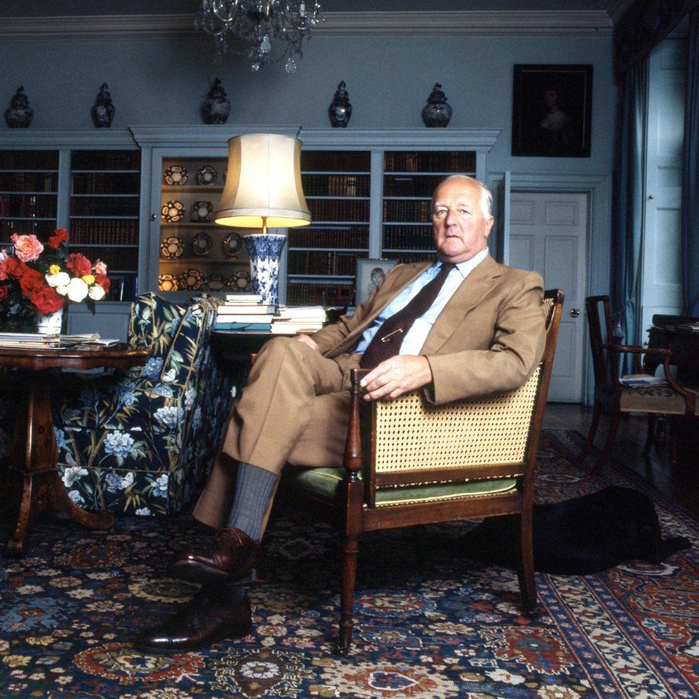 A British Duke sitting at home in a brown suit, white shirt, and burgundy tie. He's surrounded by fancy rugs, lamps, and wood furniture. 