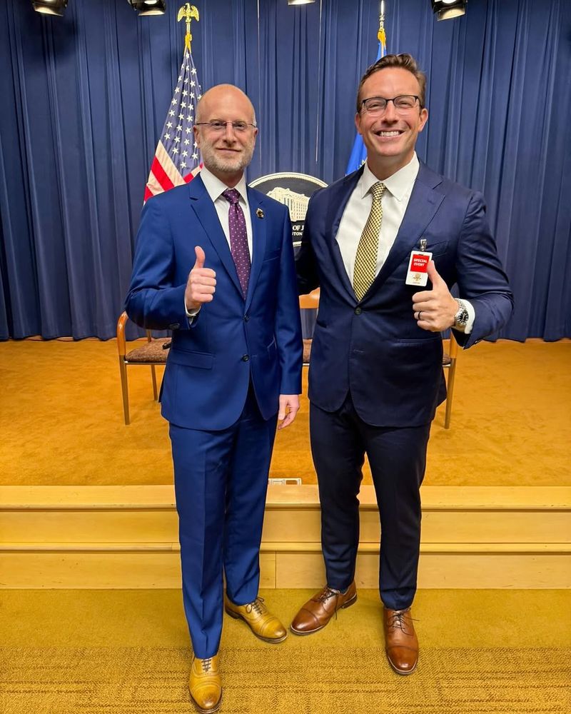 Brendan Carr and Benny Johnson. Both wearing blue suits with tan shoes. Carr's suit is mid-blue. He has paired it with blonde shoes. Johnson is wearing a navy suit with walnut colored shoes. 