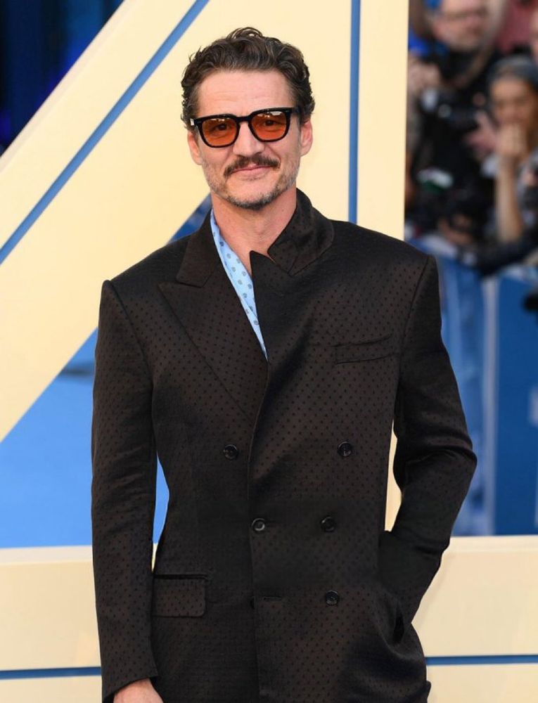 Pedro Pascal wears a dark colored double-breasted suit. One of the lapels is flipped up. 