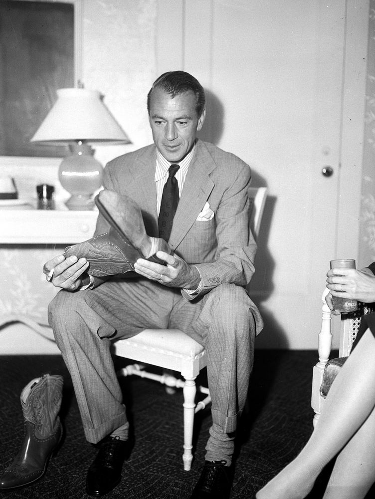 Gary Cooper is sitting down while wearing a striped shirt, single-breasted suit, and black dress shoes. His tie is worn with a four-in-hand knot. 