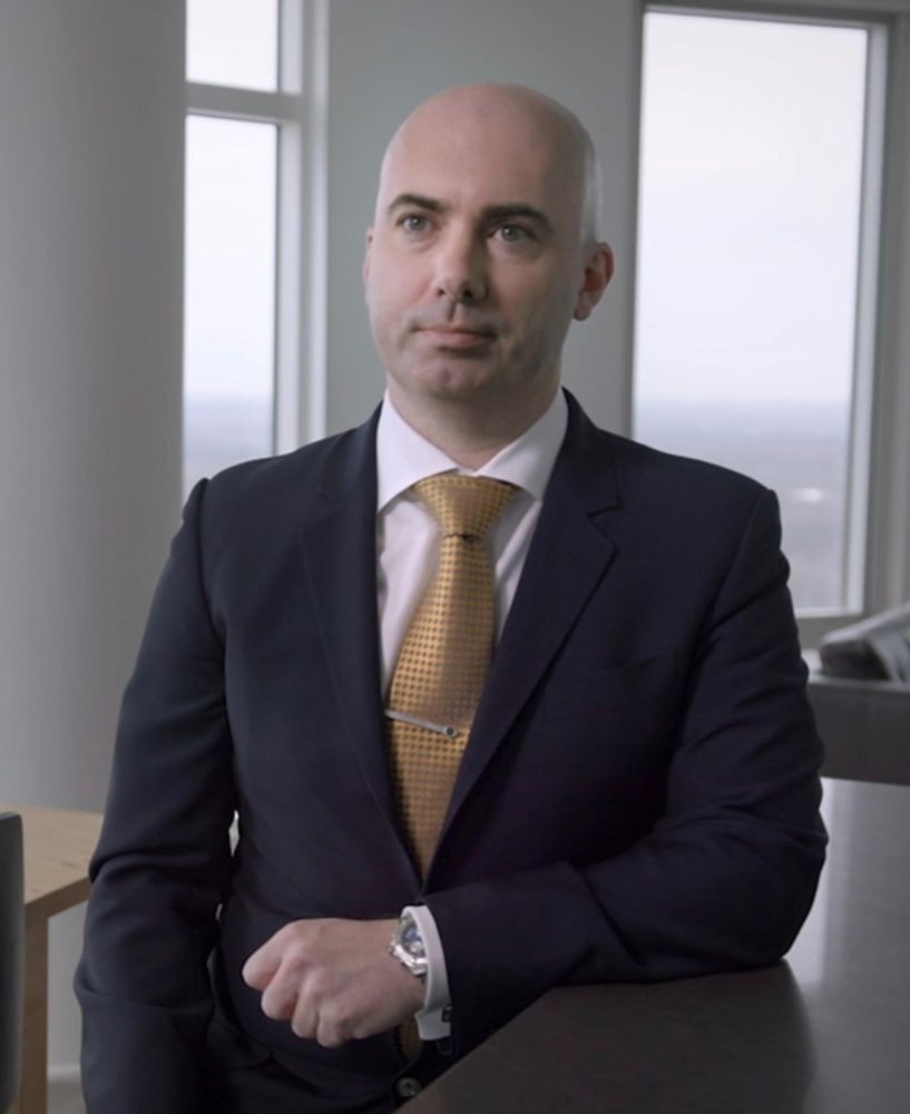 Matt Braynard wearing a navy suit with a white French cuff shirt and gold colored tie. He has a giant Windsor knot and a metal sports watch. 