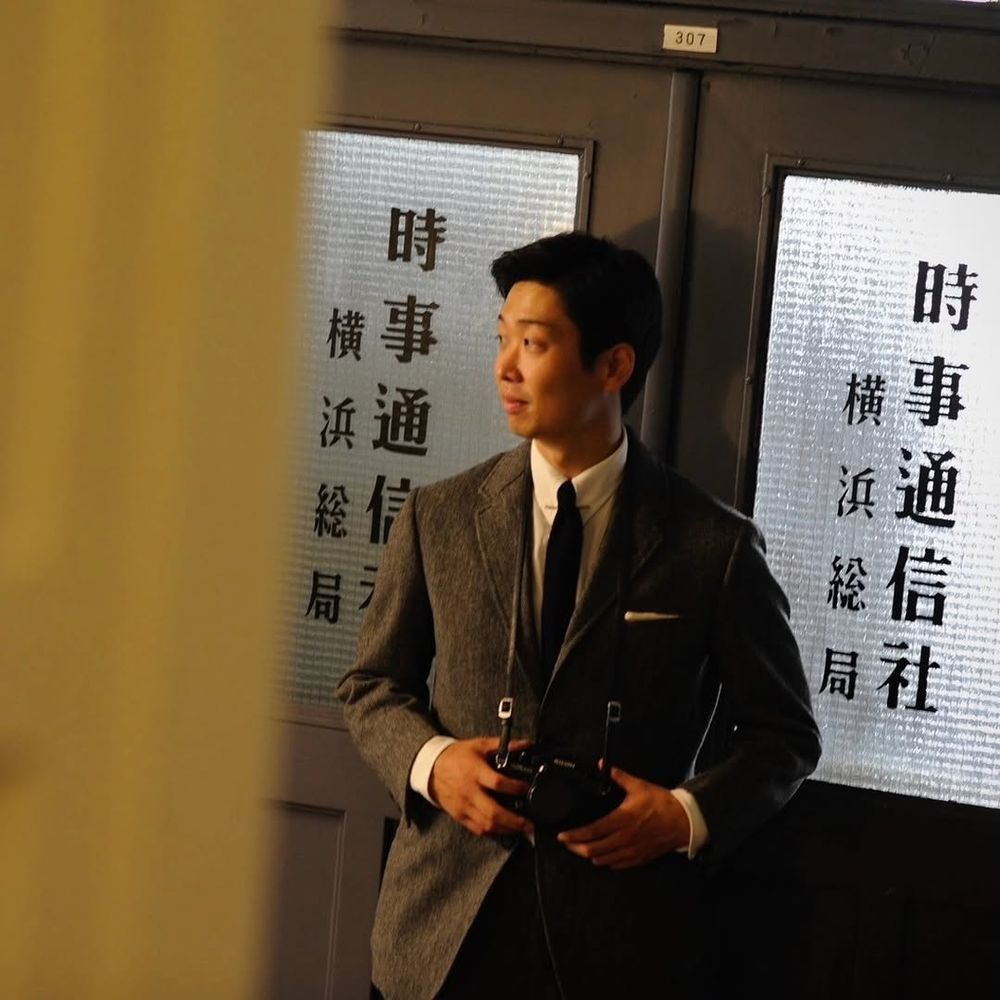 A Japanese man wearing a grey tweed sport coat, white shirt, and black knit tie