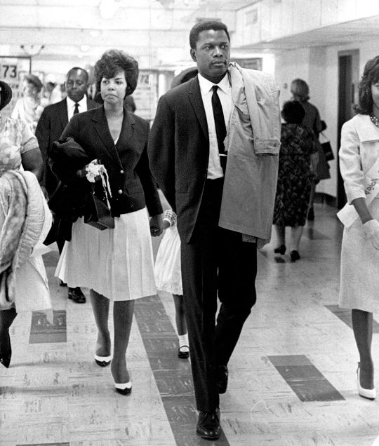 Sidney Poitier is wearing a dark single-breasted suit with a white dress shirt. His tie is worn with a four-in-hand knot. 