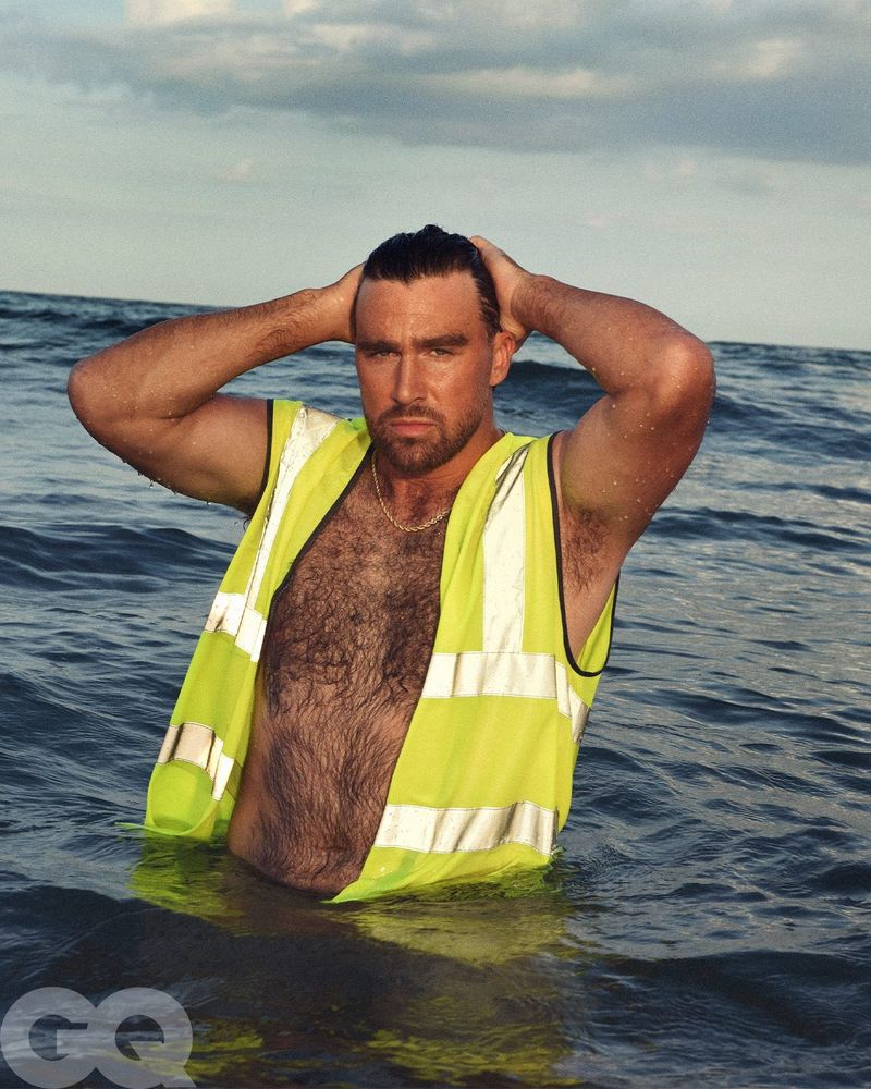 Travis Kelce in GQ. He's in the ocean and only wearing a CAT work vest and gold necklace. He's smoothing his hair back.