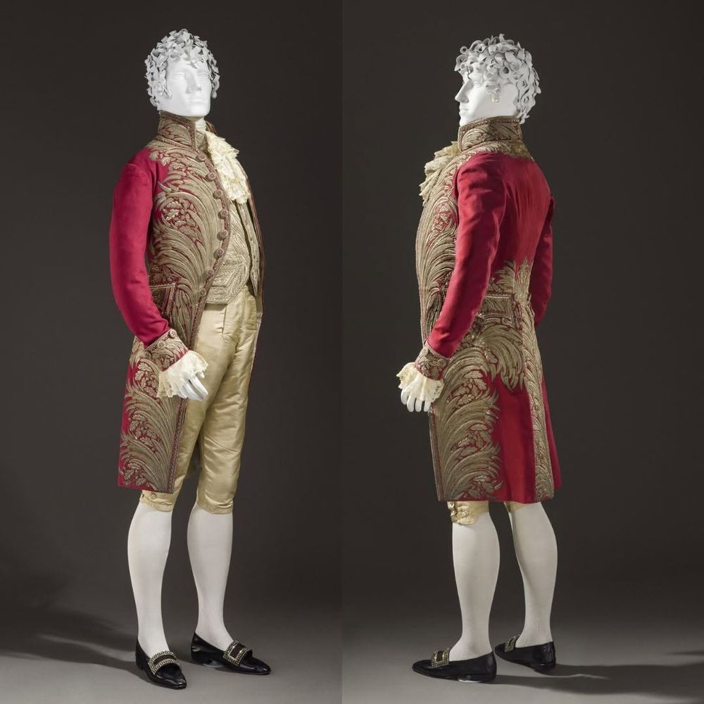A photo of British court dress in the early 1800s. The outfit consists of a red coat with brilliant gold detailing, gold colored breeches, gold colored vest, white cravat, white stockings, and black buckled shoes. 