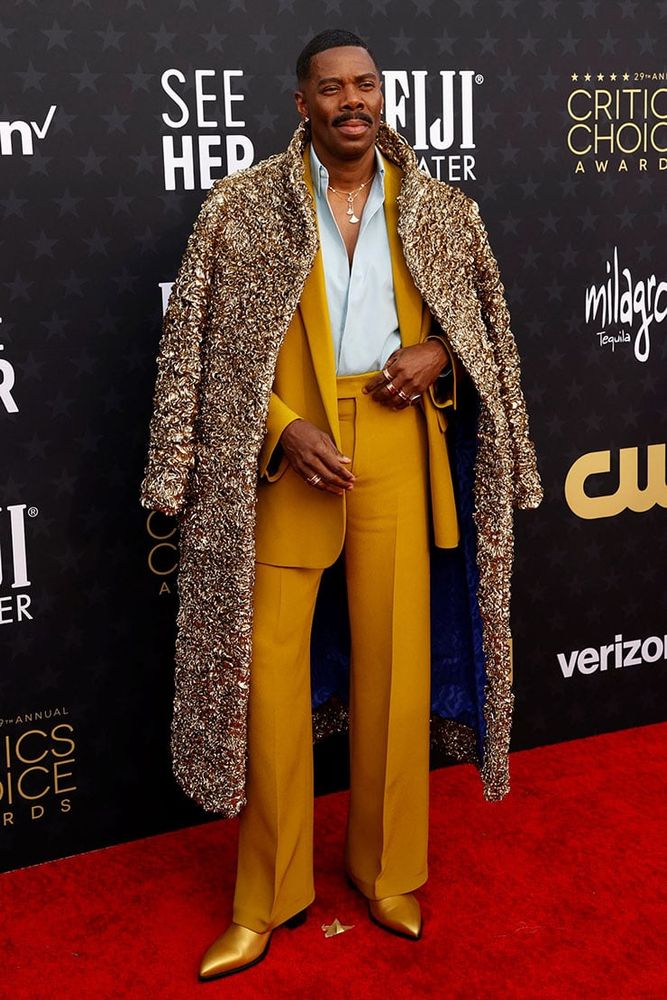 Colman Domingo wearing a gold colored Valentino suit with a light blue dress shirt, gold colored shoes, and a textured, gold-colored overcoat. 