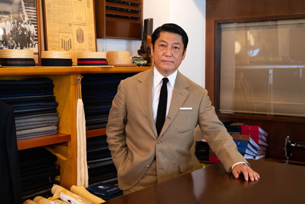 A Japanese man wearing a brown suit, white shirt, and black knit tie