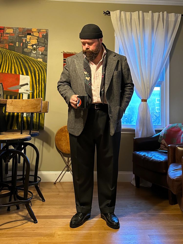 A man wearing a grey Donegal tweed sport coat with charcoal wool trousers, black Norwegian split-toes, pink-striped oxford cloth button-down shirt, brown scarf, and brown beanie. 