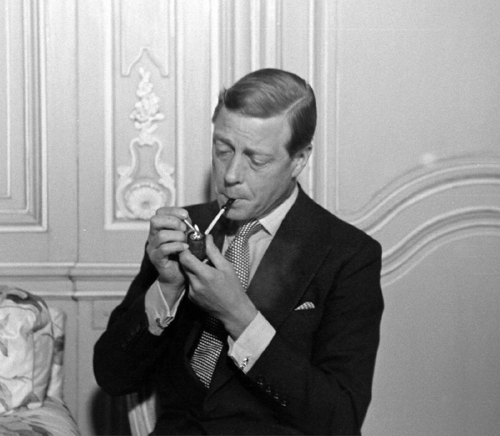 The Duke of Windsor wearing a dark suit, French cuff shirt, and necktie with a fat tie knot. He's smoking a pipe. 
