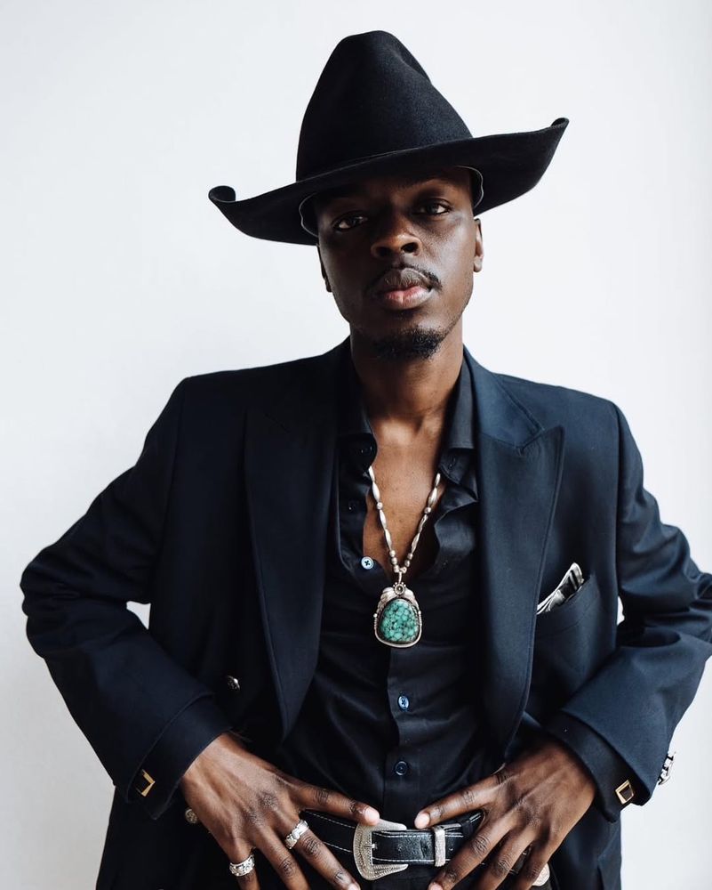 A man wears a black suit with black silk shirt, some jewelry, and a black cowboy hat