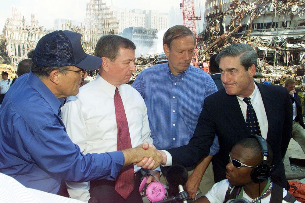 Robert Mueller shakes hands with Rudy Giuliani on 9/11. Smoldering buildings are behind them. Mueller is wearing a dark suit, white shirt, and dark tie. 