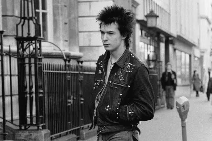 Sid Vicious on the street wearing a studded leather jacket, jeans, and a chunky necklace. He has slightly spiked hair. 