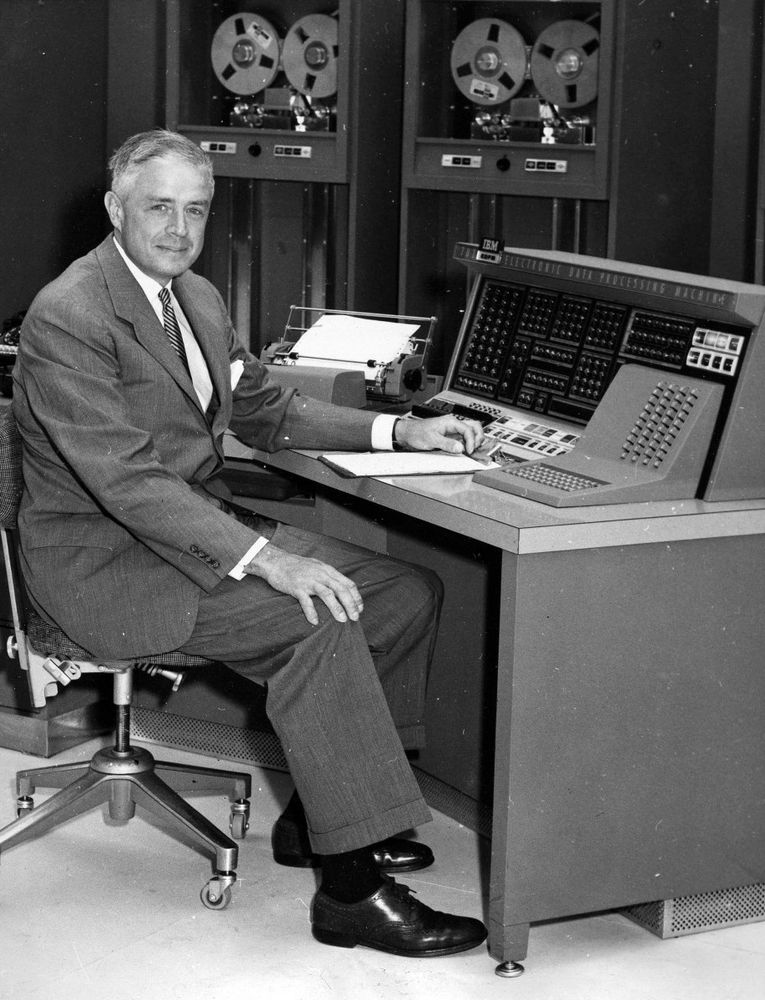 Thomas Watson, Jr. of IBM wearing a dark suit, white shirt, dark tie, and black oxford shoes. He's sitting in front of a computer. 