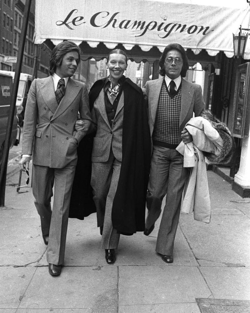 Valentino Garavani, Giancarlo Giammetti and Francoise de la Renta outside Orsini's on February 29, 1972 in New York. Giammetti is wearing a 6x2 double breasted suit with long spearpoint collar and tie. De la Renta  is wearing a single-breasted suit with a cardigan, scarf, and long overcoat. Garavani is wearing a single-breasted suit with a checked sweater vest, spearpoint collar, and tie. 