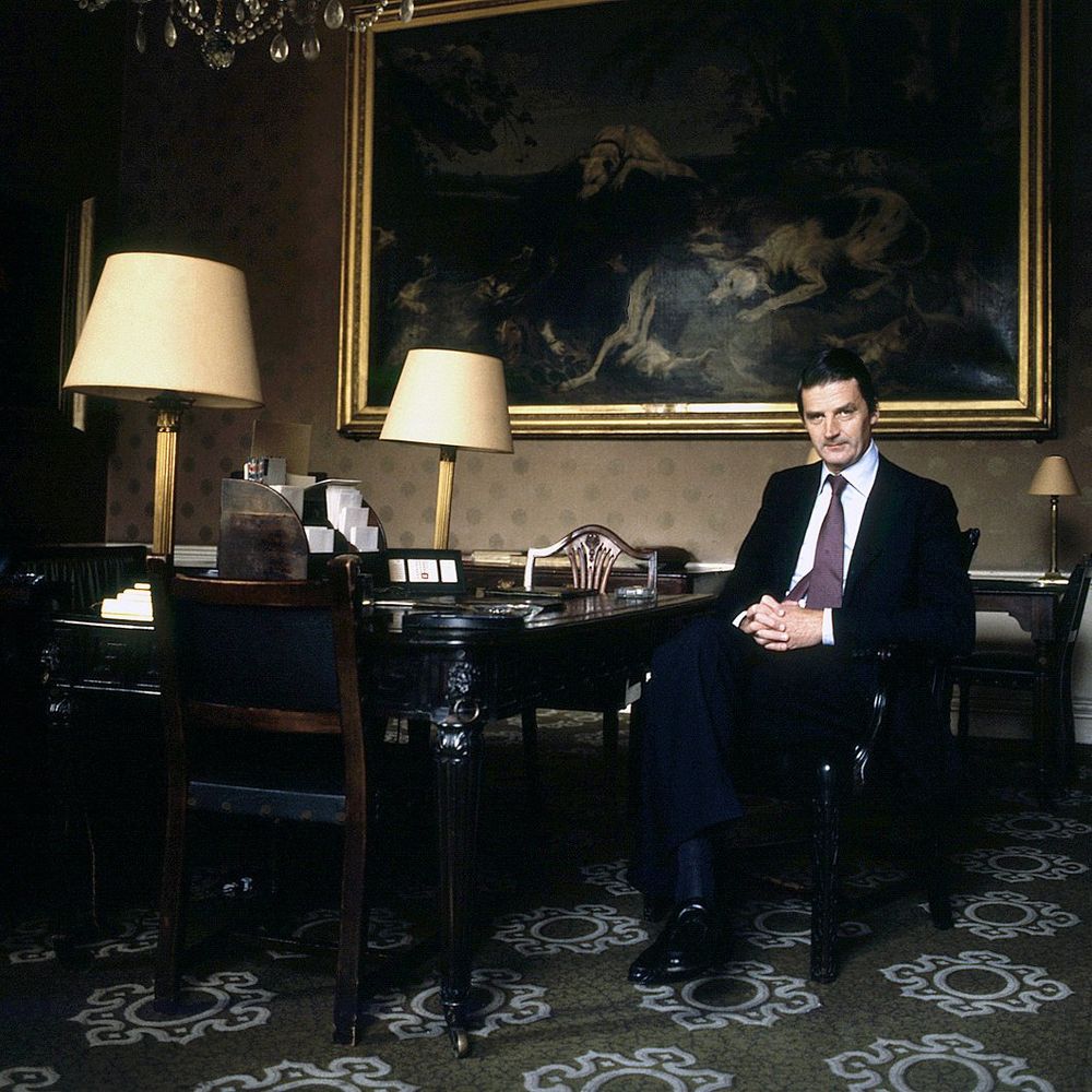 A British duke sitting at home in a dark worsted suit, white shirt, and black oxfords. He's sitting at a fancy wood desk with fancy lamps and a big painting in the back. 