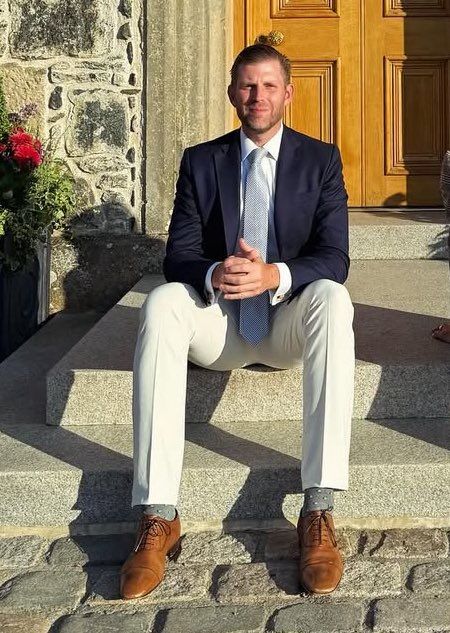 Eric Trump wearing a navy sport coat with a white shirt, silver colored tie, off-white trousers, and tan oxfords. 