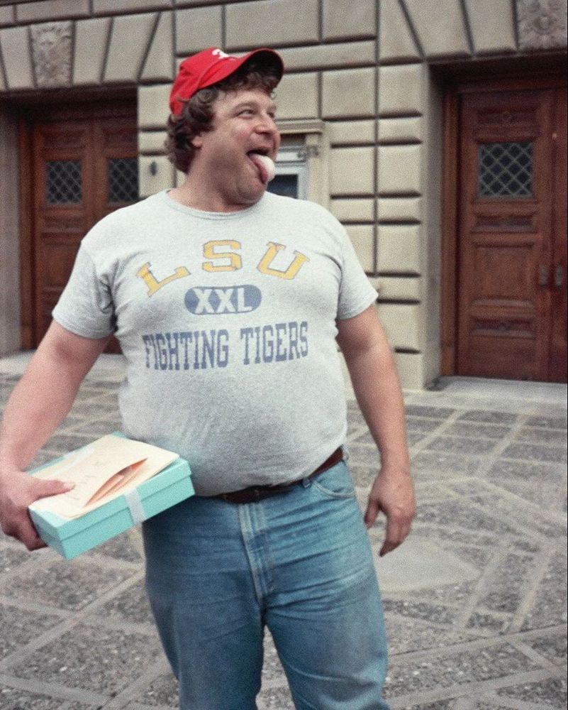 John Goodman in a grey LSU Fighting Tigers t-shirt, blue jeans, and a red baseball cap