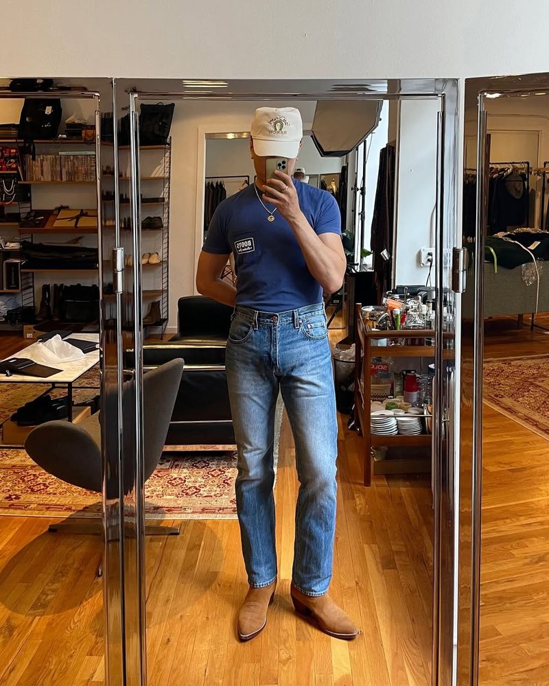 A man wears mid-rise, bootcut jeans with suede cowboy boots, tight t-shirt, and a trucker cap