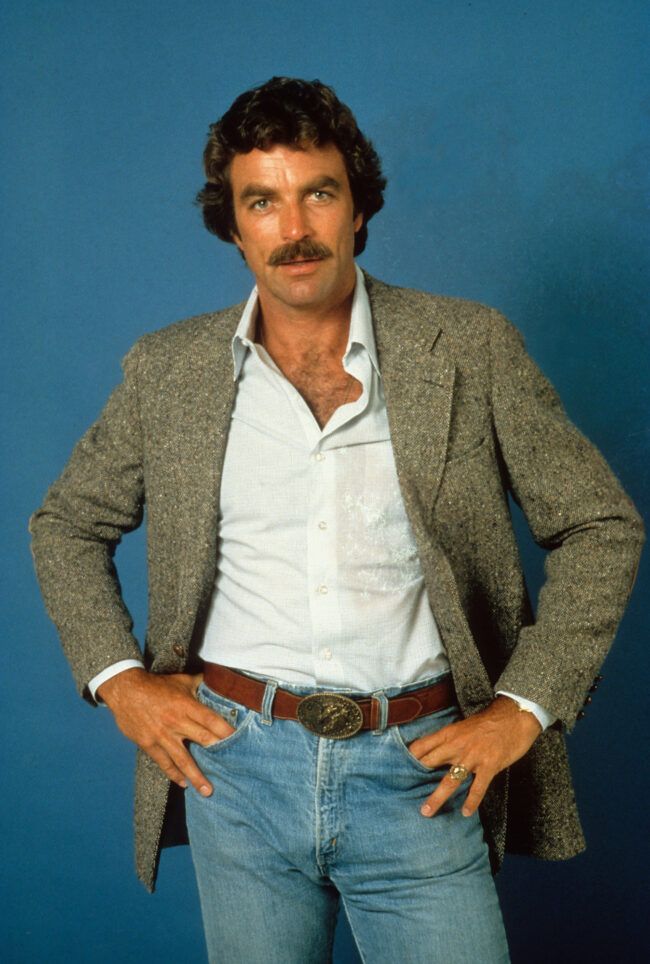 Tom Selleck wearing a grey Donegal tweed with light blue jeans, a brown Western buckle belt, and white dress shirt. 